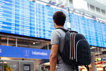 Young,Man,With,Backpack,In,Airport,Near,Flight,Timetable