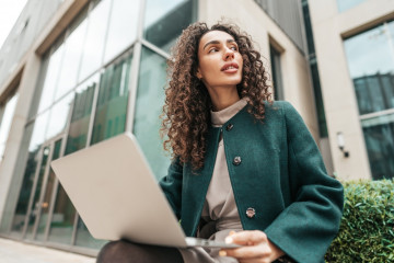 Young,Woman,Using,Laptop,While,Sitting,By,The,Building,Outdoors.